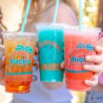Three hands hold up a variety of colorful Bahama Buck's Red Bull Infusions in a toast on a sunny day. The drinks shown include a layered orange and red infusion, a bright teal one, and a pink one.