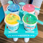 A person holds a Bahama Buck's four-cup drink carrier that reads 'Flavor Your Life.' The carrier holds four drinks, each with a colorful, decorative lid that looks like a paper umbrella and has a spoon tucked into it.