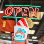 A hand holds up a red-and-white striped Bahama Buck's shaved ice in front of a window with a glowing red neon 'OPEN' sign in the background.