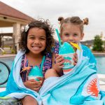 Two smiling young girls are wrapped together in a shark-themed towel while sitting by a swimming pool. They are both holding blue Bahama Buck's Shark Attack shaved ice treats.