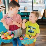 Two young boys smile at each other inside a Bahama Buck's shop while holding up Easter baskets filled with colorful eggs and packets of Bahama Buck's Sno Pops.