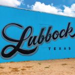 A large, popular mural on a bright blue wall that reads 'Lubbock' in a large, black cursive script, with 'TEXAS' in smaller capital letters below it.