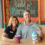 A man and a woman, both wearing Bahama Buck's polo shirts, smile for a portrait while sitting at a table inside their shop. In front of them are a blue shaved ice and a magenta smoothie, with a 'Taste of Paradise' sign on the wall behind them.