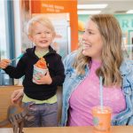 A candid photo of a mother and her young son enjoying treats at Bahama Buck's. The mother smiles lovingly at her son, who makes a funny, scrunched-up face while holding his shaved ice. A toy dinosaur sits on the table beside them.