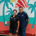 Lavern Pena and her husband stand side-by-side, smiling in front of their Bahama Buck's trailer. Both are wearing Bahama Buck's polo shirts, and the trailer behind them features a large, colorful graphic of a shaved ice and palm trees.