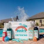 A Bahama Buck's catering pack is set up on the edge of a swimming pool with a large splash of water in the background. The display includes a cooler of shaved ice, several bottles of flavor syrup including Shark Attack and Cherry, cups, and spoons.