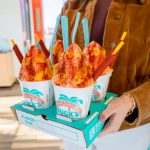 A person carries a Bahama Buck's drink tray holding four Mango shaved ice treats, each topped with a red chili-lime seasoning and garnished with a tamarind candy straw.