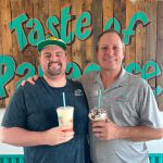 Two men smile for a photo inside a Bahama Buck's, standing in front of a wooden wall with the words 'Taste of Paradise.' Each man is holding a different Bahama Buck's specialty drink.