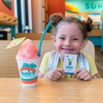 A smiling young girl with her hair in two space buns sits at a table inside a Bahama Buck's. She proudly holds up a reward card for a 'FREE' treat, with a large pink shaved ice on the table next to her.