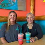 Jessica Pool and Lolli Ulibarri sit side-by-side at a table inside a Bahama Buck's shop, smiling for the camera. On the table in front of them are two colorful Bahama Buck's drinks.