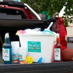 A tailgate scene featuring a Bahama Buck's Paradise Party Pack set up on the back of a pickup truck. The display includes a cooler of shaved ice, bottles of Blue Cotton Candy and Fanta Orange syrup, cups, umbrellas, and a football.