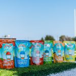 A lineup of six different flavors of Bahama Buck's SnoBlast pouches standing in a row on the white line of a green football field. The flavors shown are Tiger's Blood, Blue Raspberry, Cherry, Extreme Sour, Mango, and Lemon.