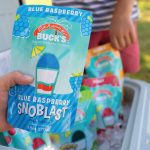 A close-up shot of a hand holding a Bahama Buck's Blue Raspberry SnoBlast pouch. In the background, an open cooler is filled with more SnoBlasts on ice.