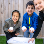 Three smiling young boys have a playful snowball fight in a backyard, grabbing perfectly round snowballs from a large styrofoam cooler filled with them.