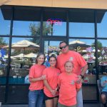Jennifer Burroughs, owner of the Bahama Buck's in Redlands, California, stands smiling with her husband, son, and daughter in front of their shop. The family is wearing matching coral-colored t-shirts, and the store windows behind them are decorated with 'Sonic the Hedgehog' characters.