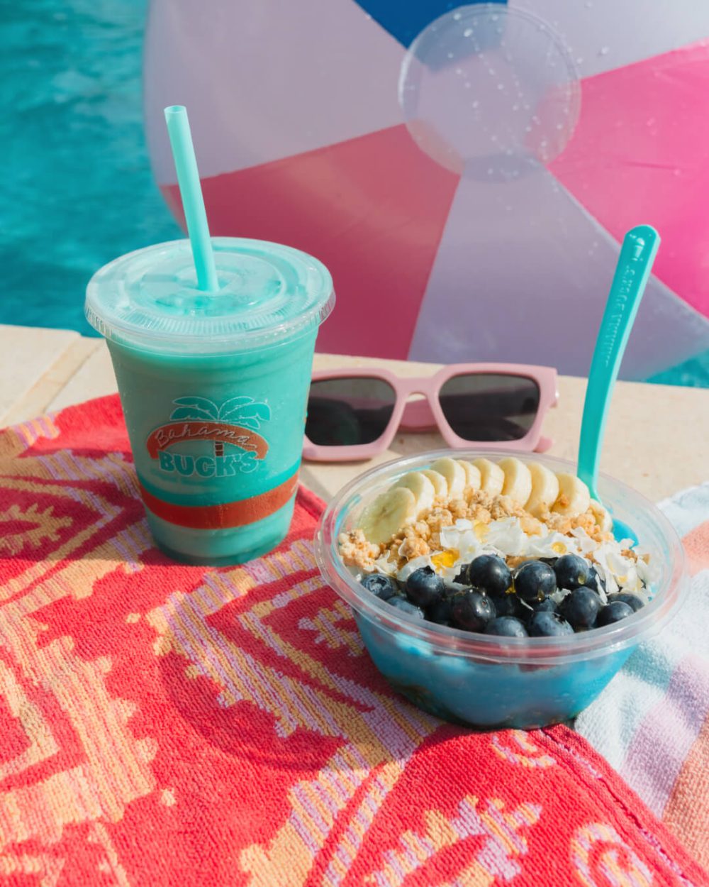 Photo of the 'Blue Colada Bowl & Smoothie' from Bahama Buck's. The bowl and smoothie sit on colorful towels next to a pool with a beach ball floating in it.