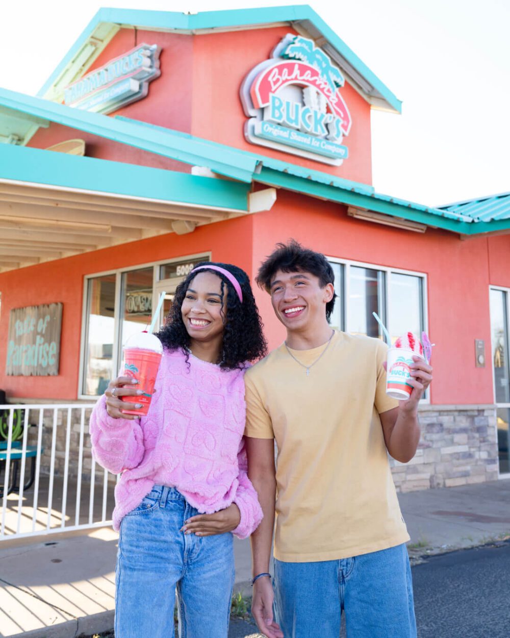 A boy and a girl in front of a Bahama Buck's shop holding a Strawberry Cheesecake Delight Sno® and Strawberry Freezecake Smoothie.